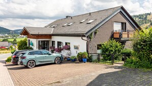 Ein zweistöckiges Haus mit Satteldach, einem Balkon im zweiten Obergeschoss und einer Auffahrt mit zwei geparkten Autos.