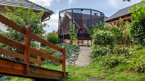 Eine Holzbrücke führt zu einem Trampolin in einem Garten mit Pflasterweg und Grünflächen.