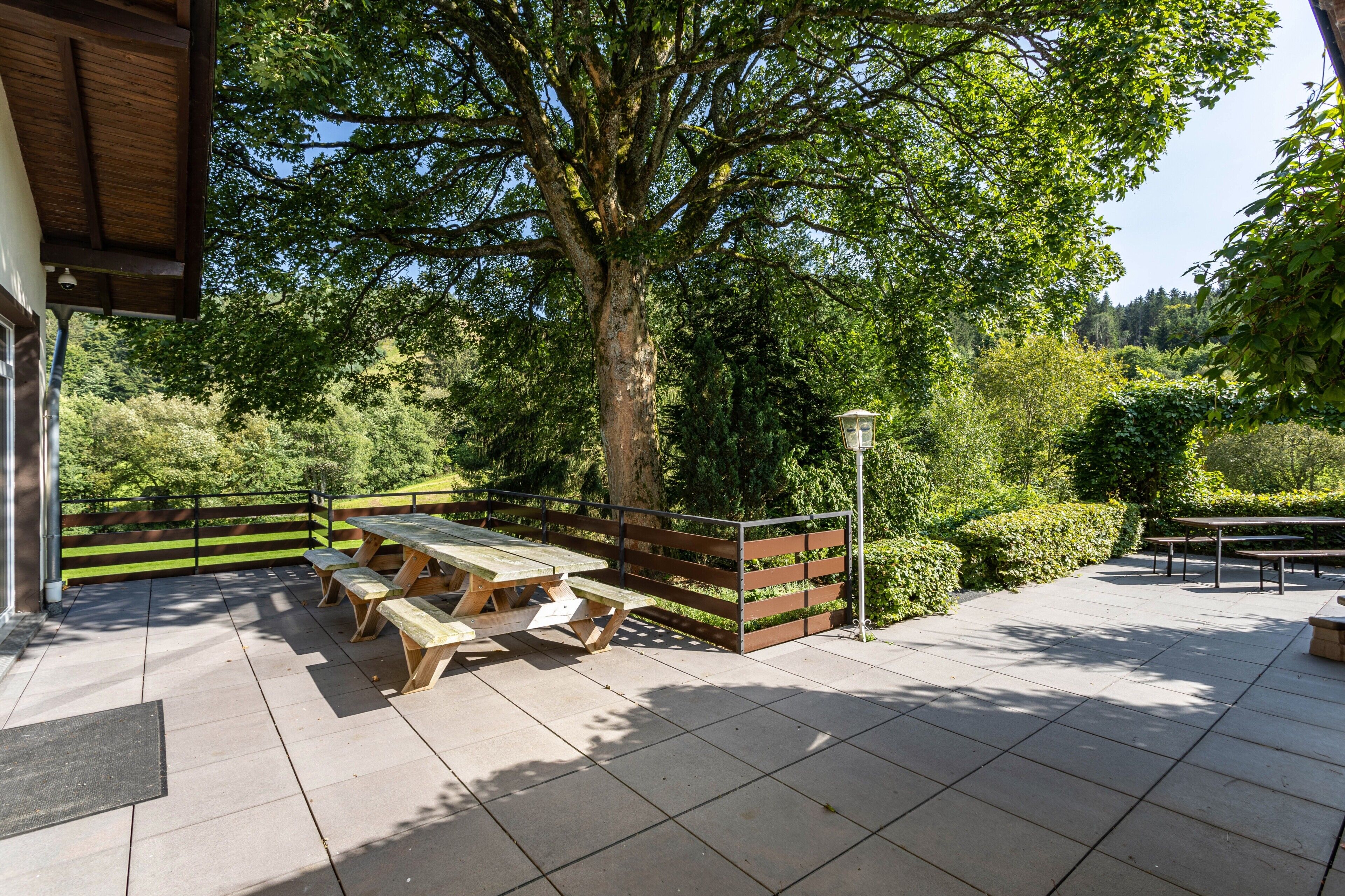 Une terrasse extérieure avec des tables de pique-nique, une clôture en bois et un grand arbre qui offre de l’ombre.