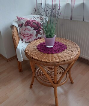 A wicker table with a potted plant and a purple doily, a chair with a floral cushion, and a window with blinds. - Apartment 'Lipperland' with Balcony and Wi-Fi (Detmold)