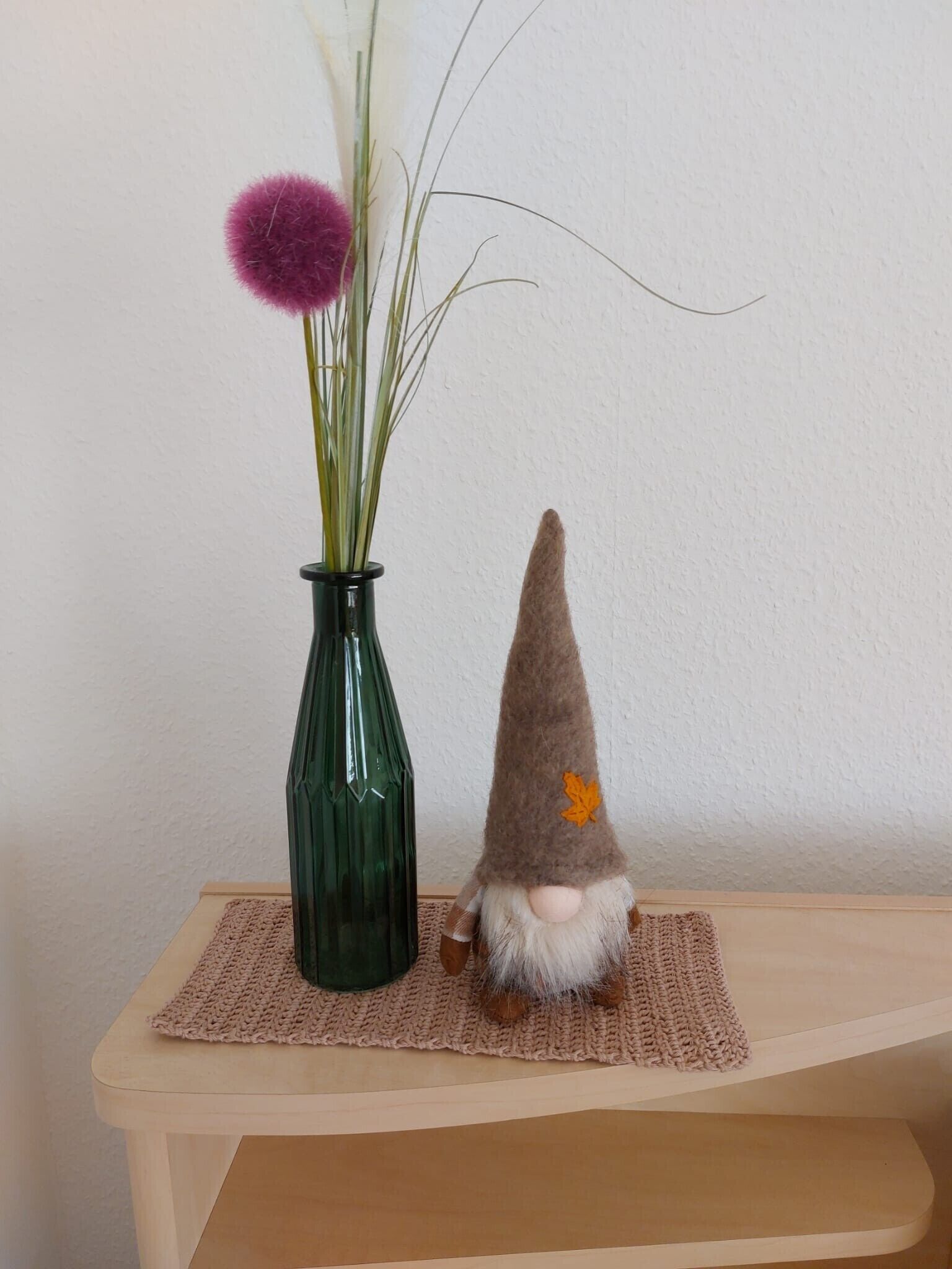 Un vase vert orné d’une fleur violette et d’une figurine de gnome, posés sur une table en bois devant un mur uni.