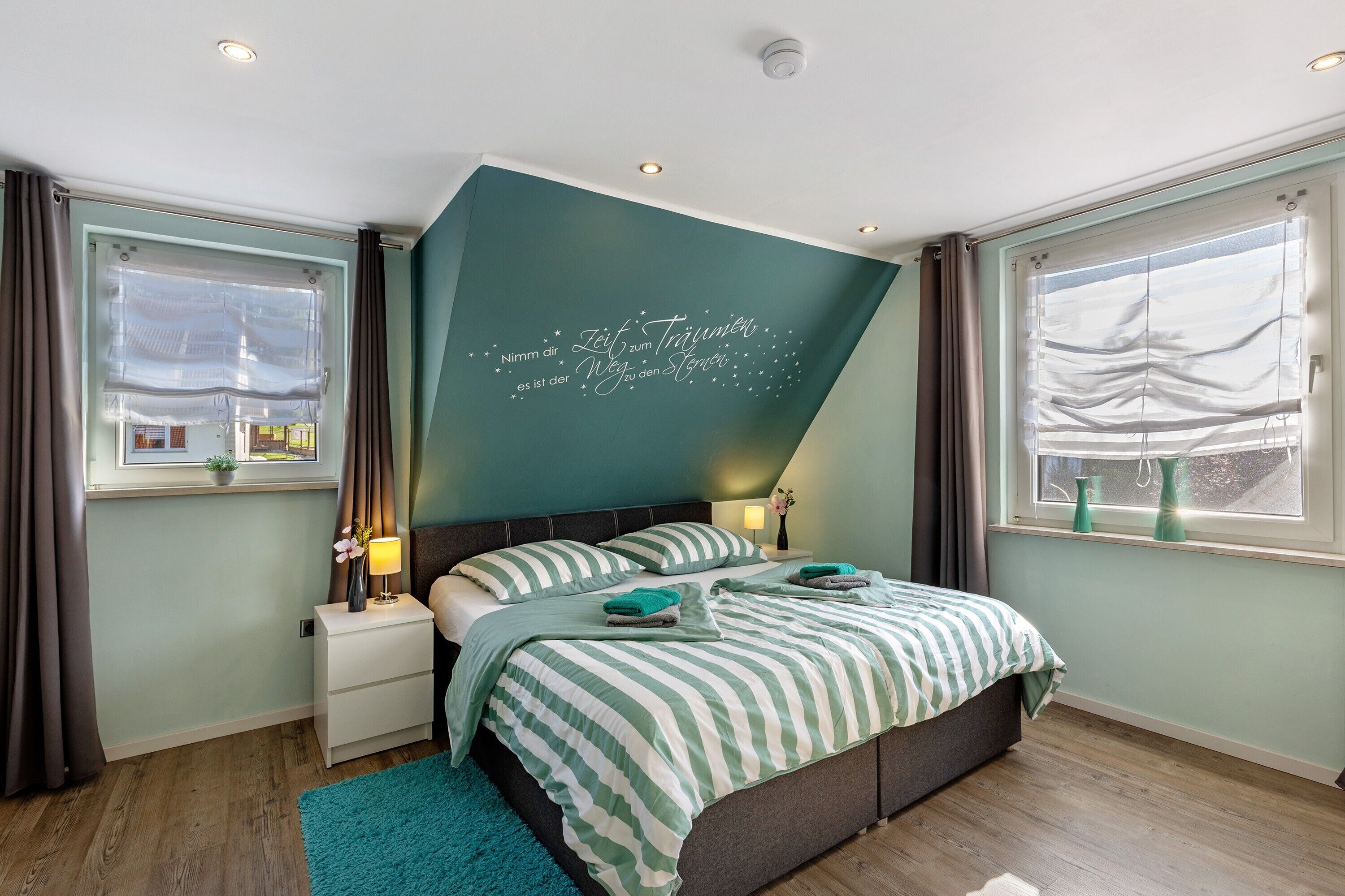 A bedroom with a bed, nightstands, a window with curtains, and a wall with a decorative phrase.