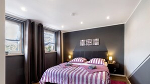 Un dormitorio moderno con una cama grande, paredes oscuras, una ventana con cortinas, una mesita de noche con lámparas y una obra de arte colgada en la pared.