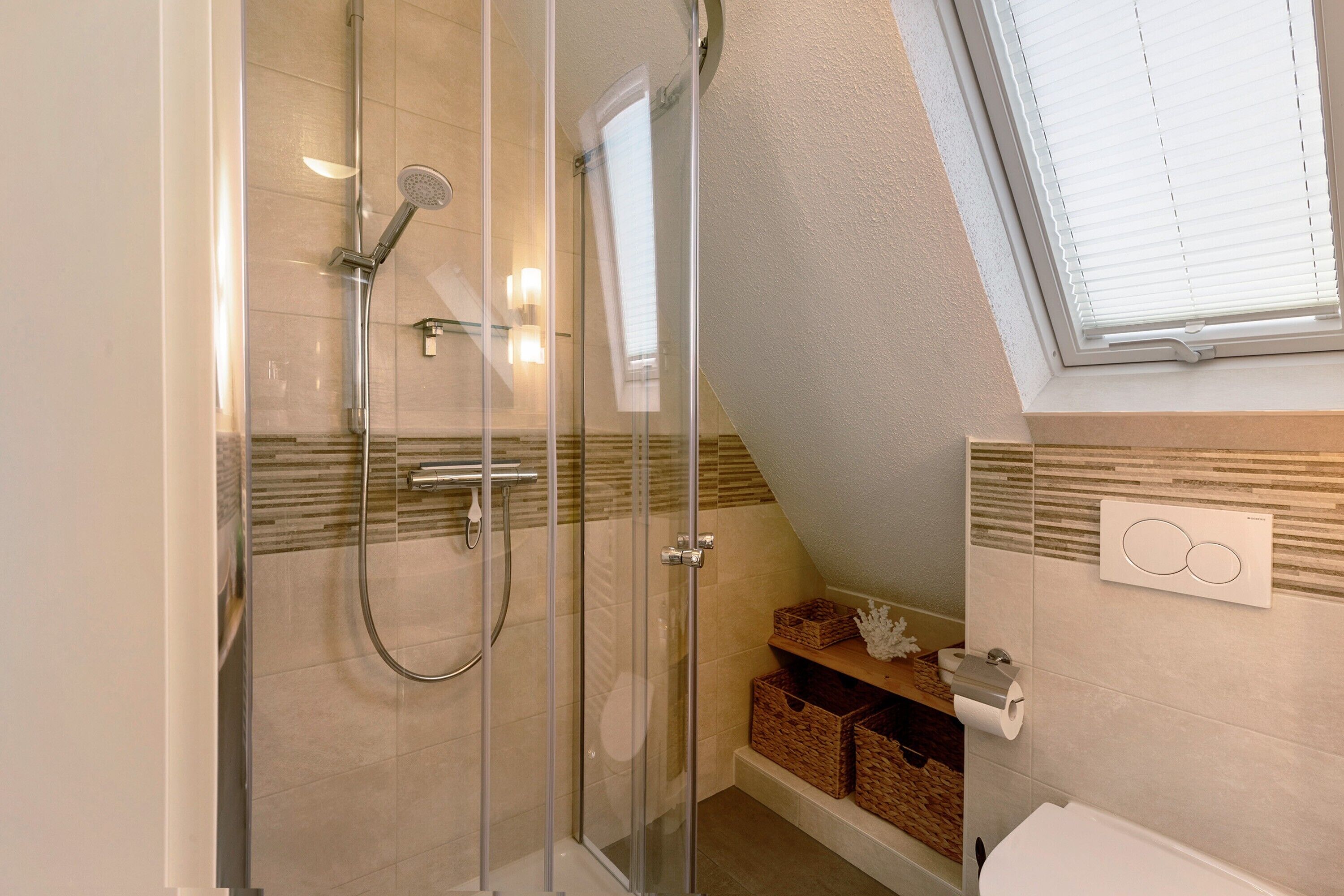 Eine Dusche mit Glastüren, eine Toilette, ein Waschbecken und ein Fenster mit Jalousien.