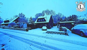 Une rue enneigée bordée de maisons en bois, une voiture garée et une autre voiture recouverte de neige.