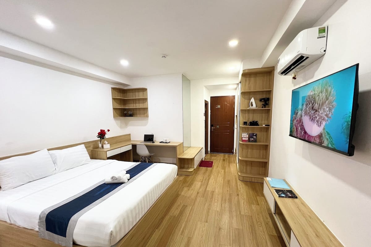 Quarto de hotel com cama, escrivaninha, cadeira, televisão e estantes.