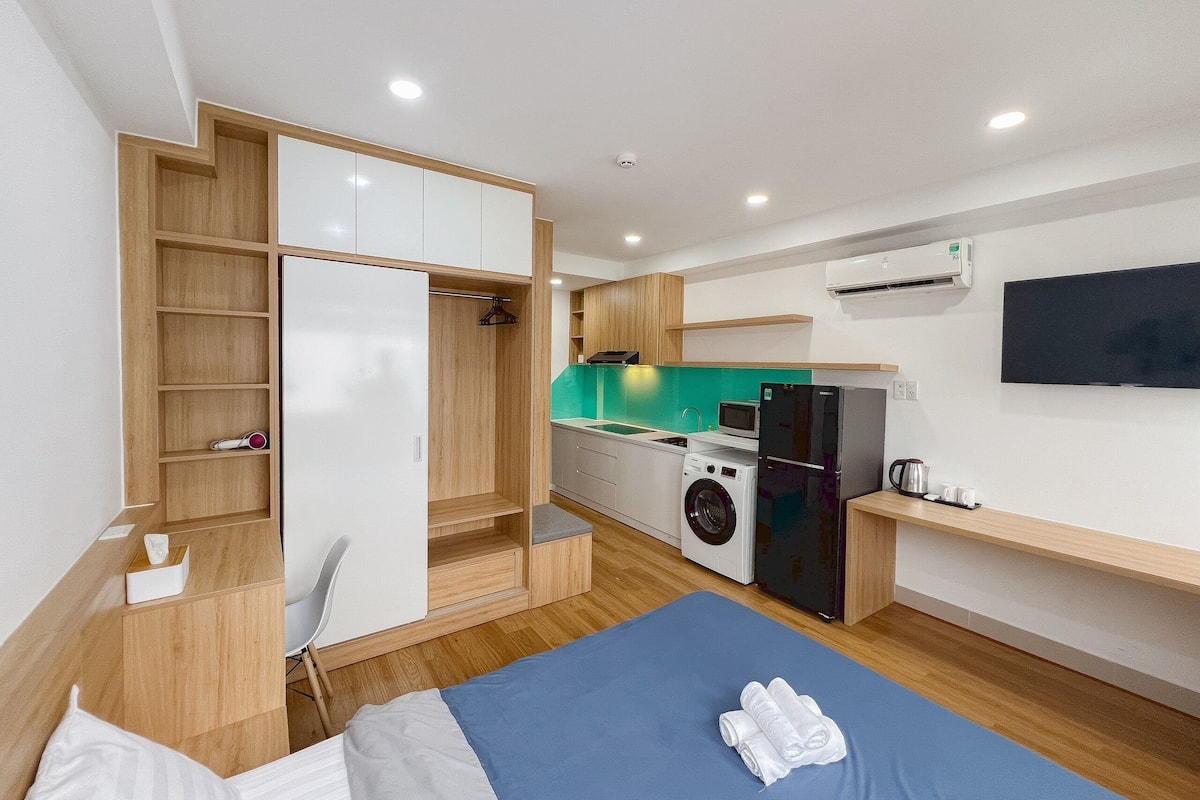 Quarto de hotel compacto com cama, pequena escrivaninha, cozinha compacta e máquina de lavar.