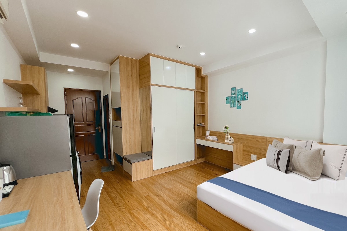 Quarto de hotel moderno com piso de madeira, cama com detalhes em azul, escrivaninha com cadeira, guarda-roupa e uma pequena cozinha.
