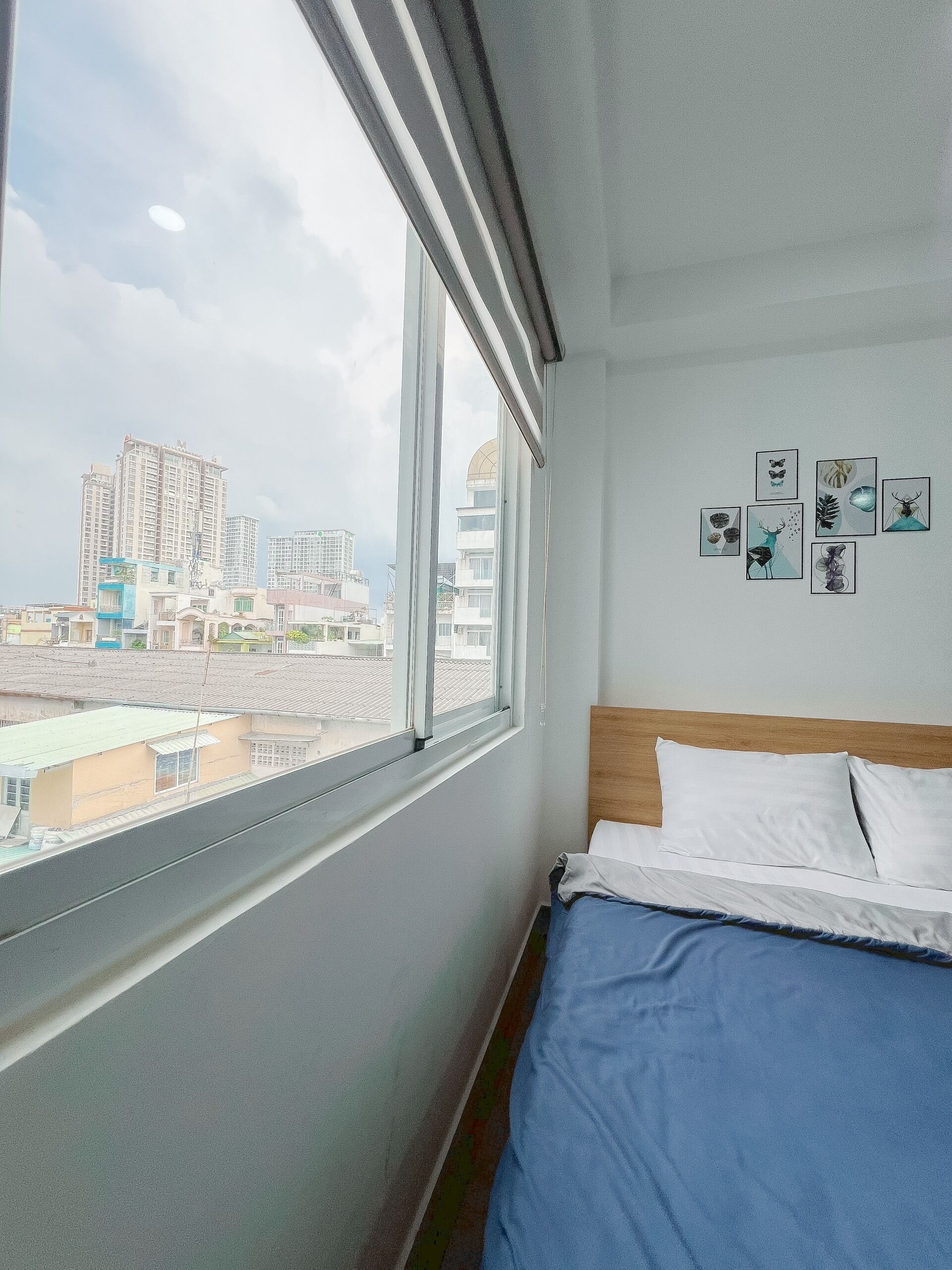 A hotel room with a large window, a bed with white and blue bedding, and several framed pictures on the wall.