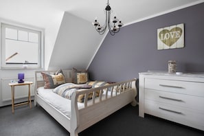 Quarto com teto inclinado, cama com várias almofadas, cômoda, luminária fixada na parede e janela com vista.