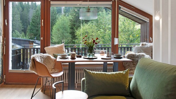 A room with a large window, a dining table, a sofa, and a hanging light fixture.