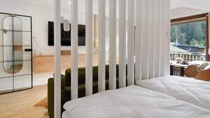 A modern hotel room with a large bed, a green armchair, a wooden cabinet, a TV, and a view of a balcony with a railing.