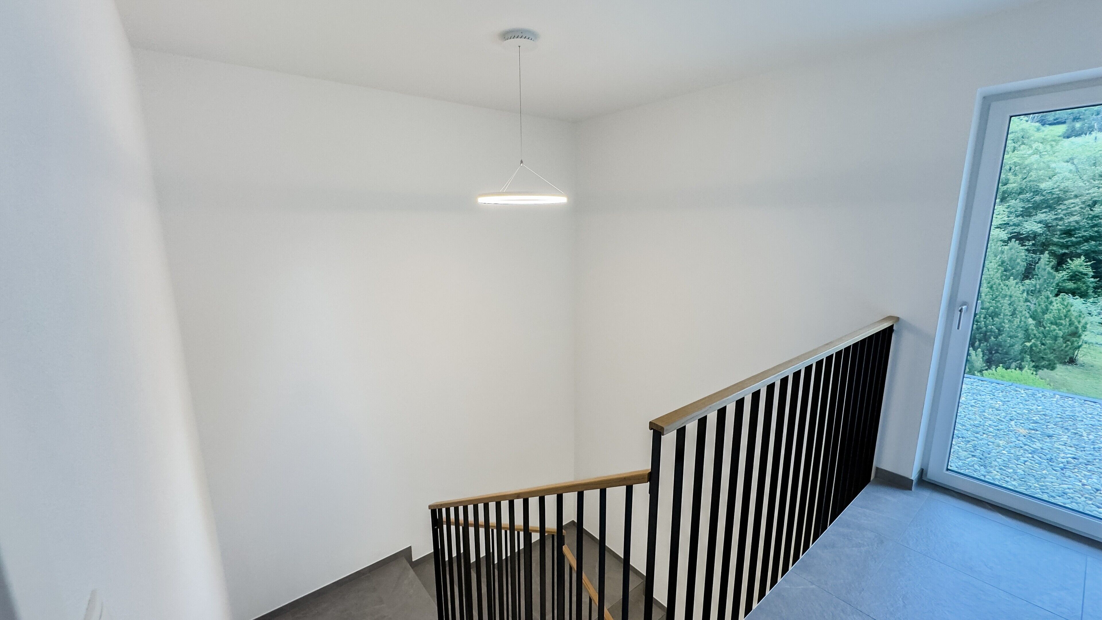 Een modern interieur met een trap, een hanglamp en uitzicht op groen door een raam.