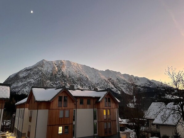 A wooden chalet with a mountain backdrop, featuring a snow-covered roof and a clear sky.