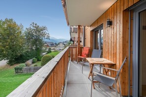 A balcony with a wooden railing, a table, two chairs, and a view of a green landscape and houses in the distance.
