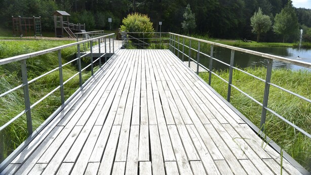 Ein hölzerner Steg mit Metallgeländern führt zu einer kleinen Holzkonstruktion, umgeben von üppigem Grün und ruhigem Wasser.