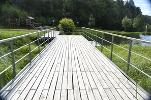 A wooden boardwalk with metal railings, leading to a small wooden structure, surrounded by lush greenery and a calm body of water.