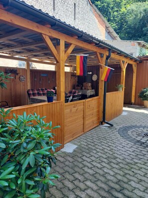 Een houten zitgedeelte buiten met een overdekt dak, een vlag en een bakstenen muur op de achtergrond.