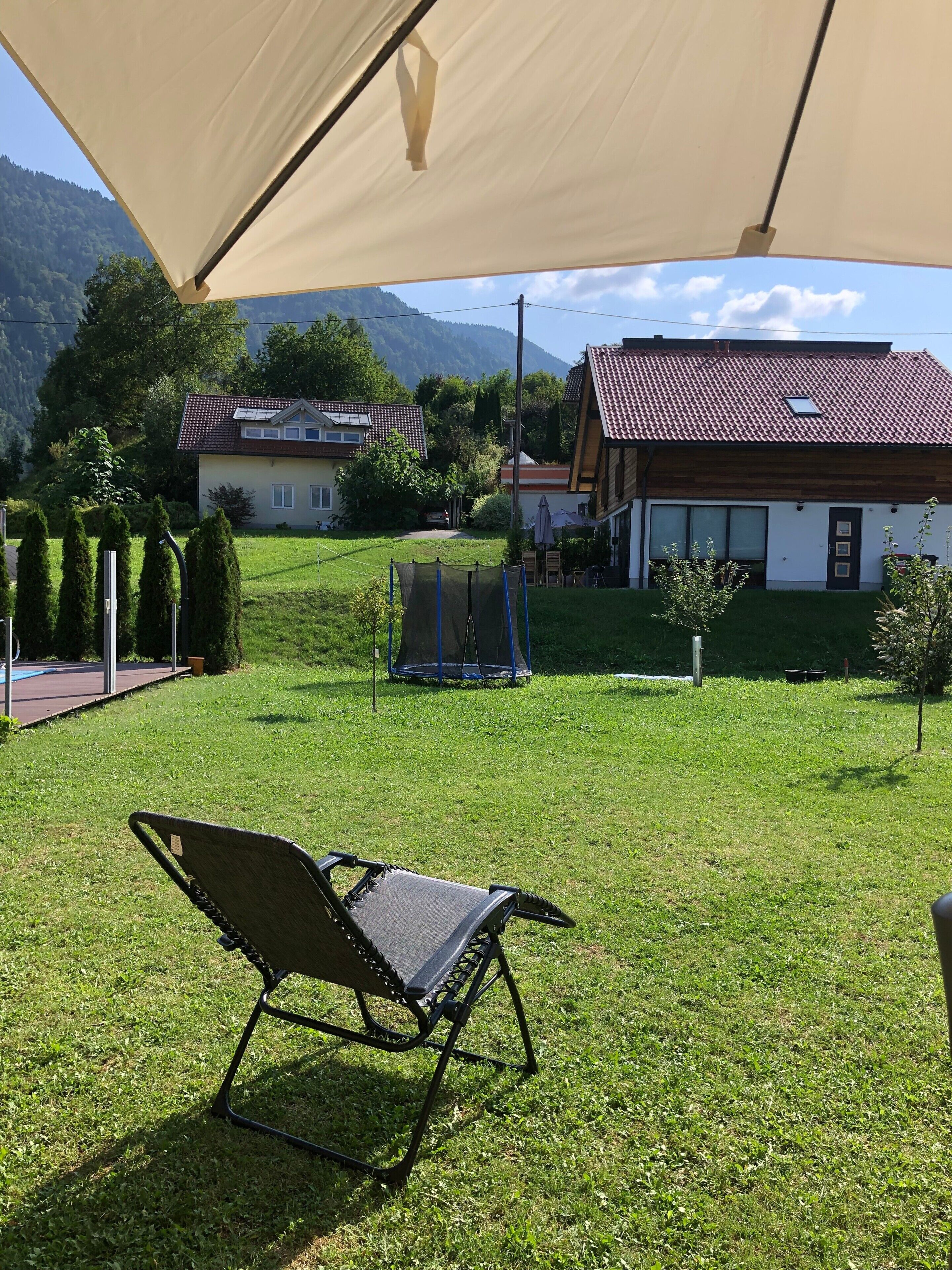 Un espace extérieur verdoyant, doté d’un coin salon ombragé, d’un trampoline et offrant une vue sur des maisons et des montagnes en arrière-plan.