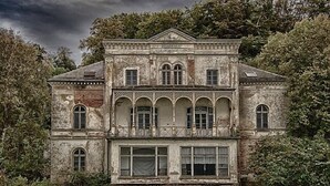 A large, historic building with multiple windows and a symmetrical facade, surrounded by lush greenery.
