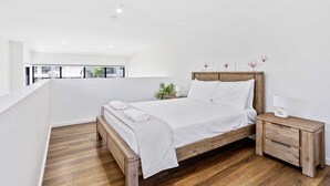 A modern bedroom with a wooden bed, white linens, a bedside table with a lamp, and a view of the outdoors.