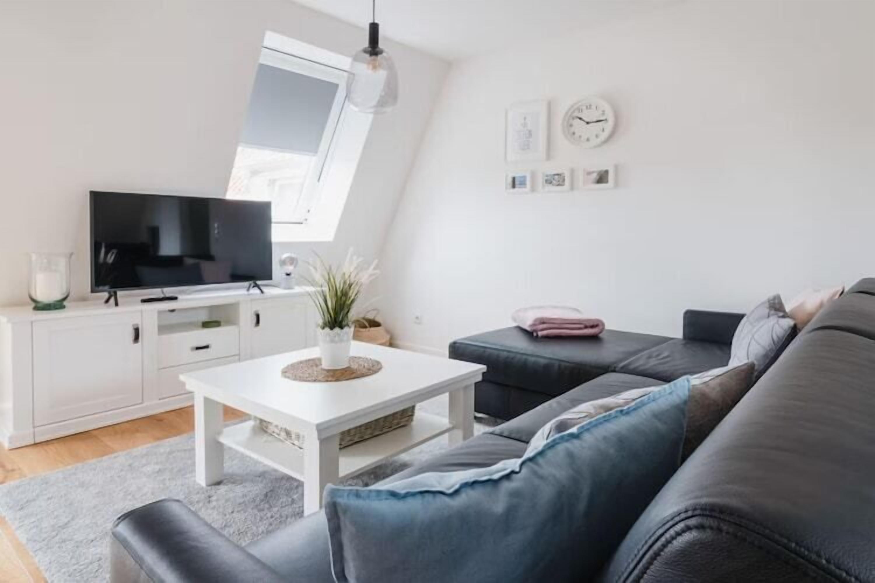 A modern living room with a flat-screen TV, white furniture, a grey sofa, and a small potted plant.