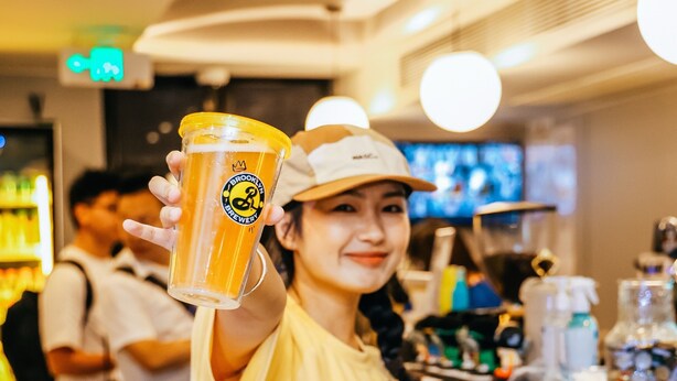 Une personne tenant un verre de bière dans un bar, avec un comptoir et des étagères en arrière-plan.