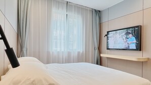 Une chambre d’hôtel avec un lit, un téléviseur fixé au mur et une fenêtre ornée de rideaux légers.