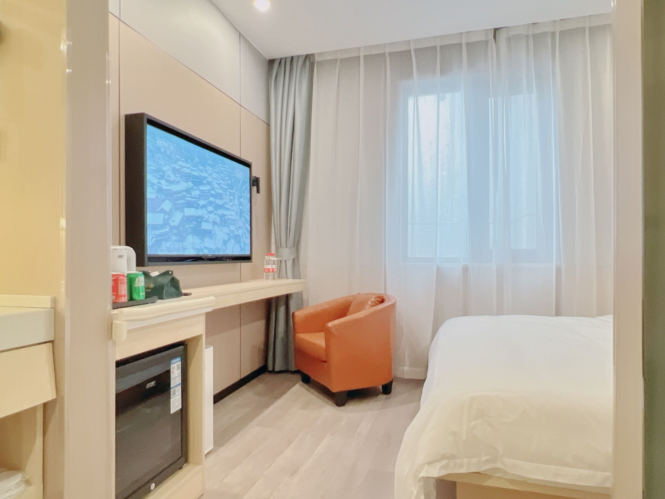 A hotel room with a bed, a TV mounted on the wall, a small desk with a computer and a bottle of water, and a window with sheer curtains.