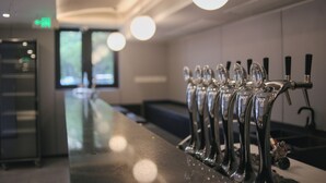 Une rangée de tireuses à bière en acier inoxydable dans un bar, avec un long comptoir et une vue sur l’extérieur grâce à de grandes fenêtres.