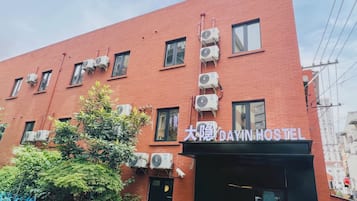 Un edificio de ladrillo rojo con múltiples unidades de aire acondicionado, un letrero que dice "DAYIN HOSTEL" y una cerca metálica azul con follaje.