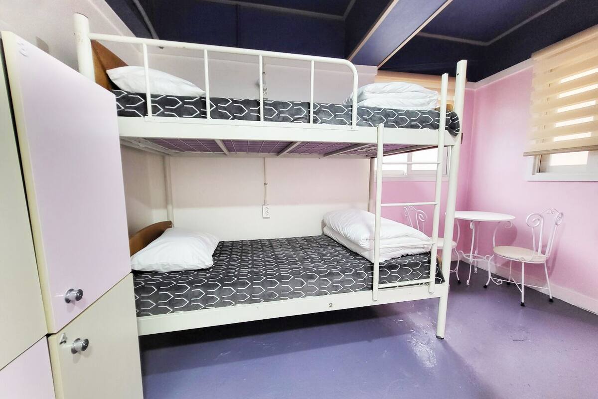 Ein Schlafraum mit Etagenbetten, einem Schreibtisch und Stuhl, einem Fenster mit Jalousien und einer rosa Wand.
