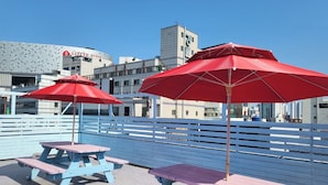Outdoor seating with red umbrellas and picnic tables, a fence, and buildings in the background. - Nampo Hill guesthouse (Busan)