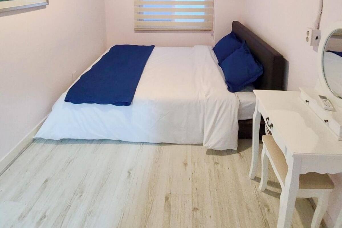 Ein kleines Schlafzimmer mit einem Bett, einem Schreibtisch, einem Stuhl, einem Fenster mit Jalousien und einer Deckenleuchte.