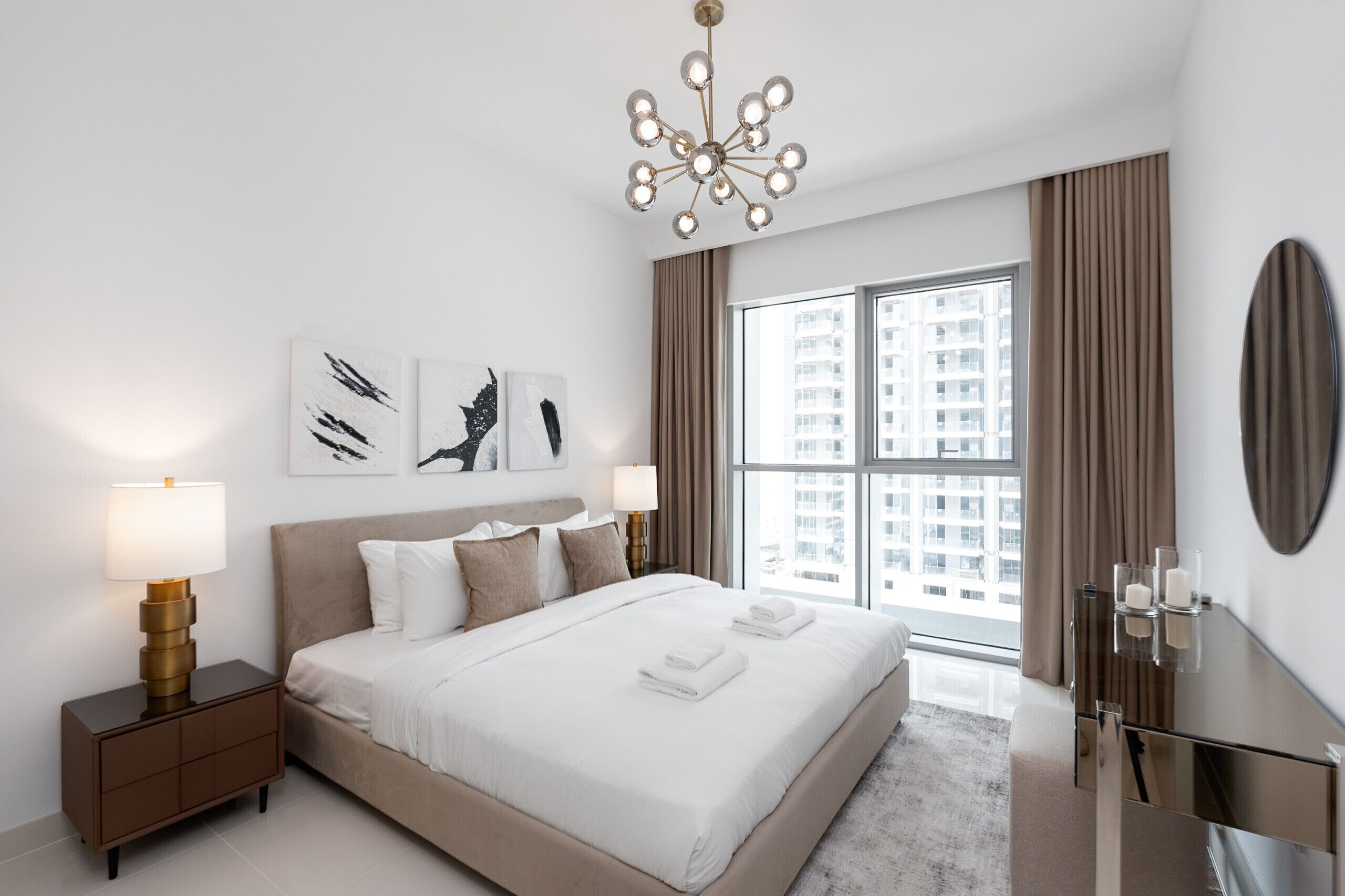 Een moderne hotelkamer met een groot bed, bedlampjes, kunst aan de muur en uitzicht op de stad door grote ramen.