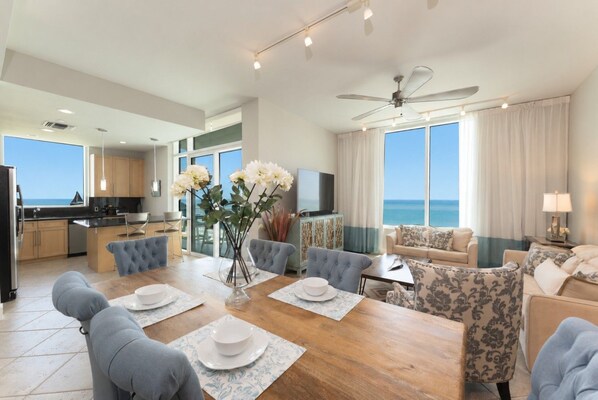 A modern hotel room with a dining area, a sofa set, a TV, and a view of the ocean.