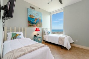 A hotel room with two beds, a painting, a TV, and a large window with a view of the sea.