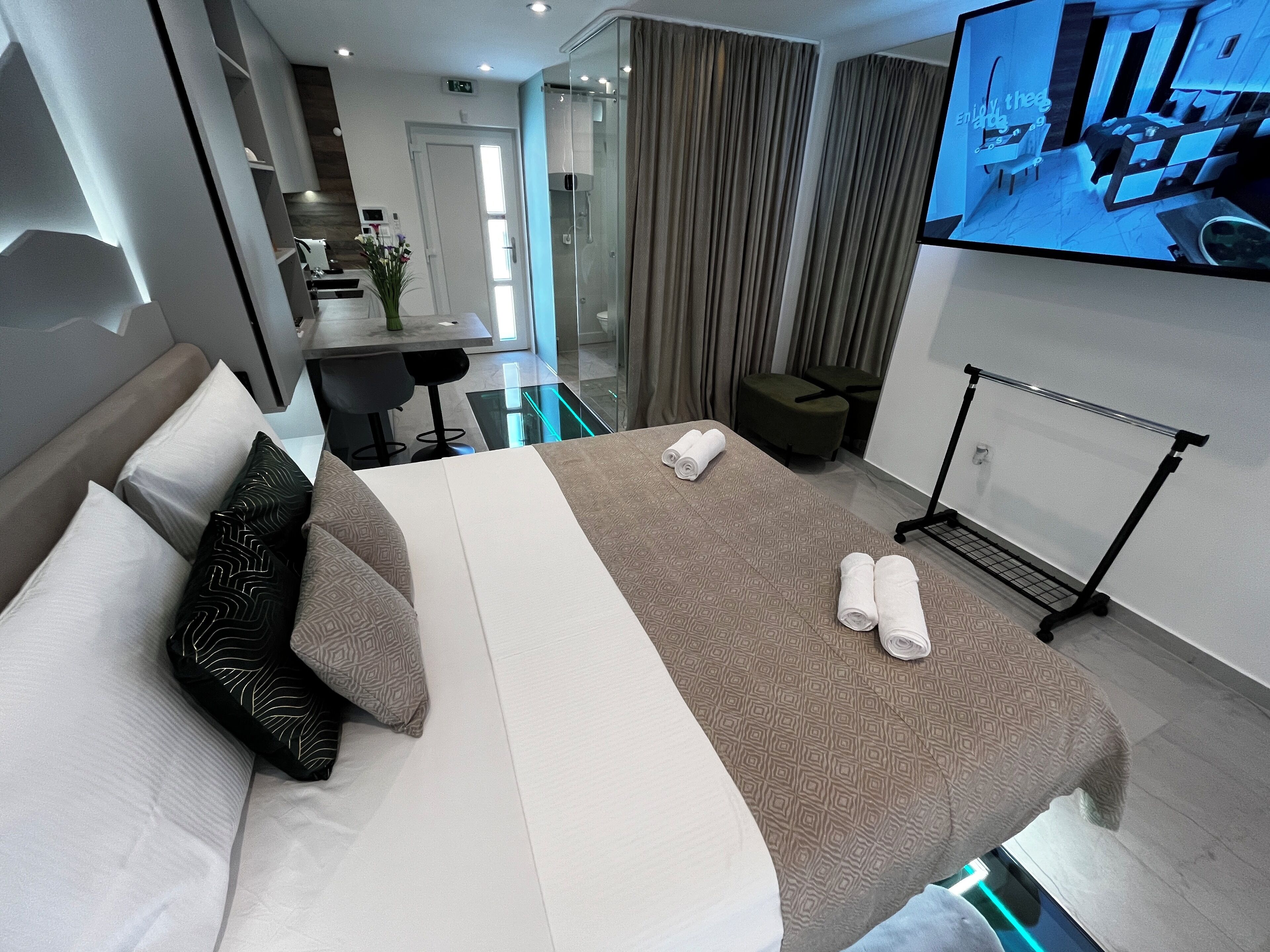 A modern hotel room with a large bed, a TV mounted on the wall, a seating area with a table and chairs, and a glass partition separating the room from the dining area.