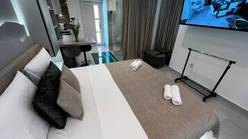 A modern hotel room with a large bed, a TV mounted on the wall, a seating area with a table and chairs, and a glass partition separating the room from the dining area.