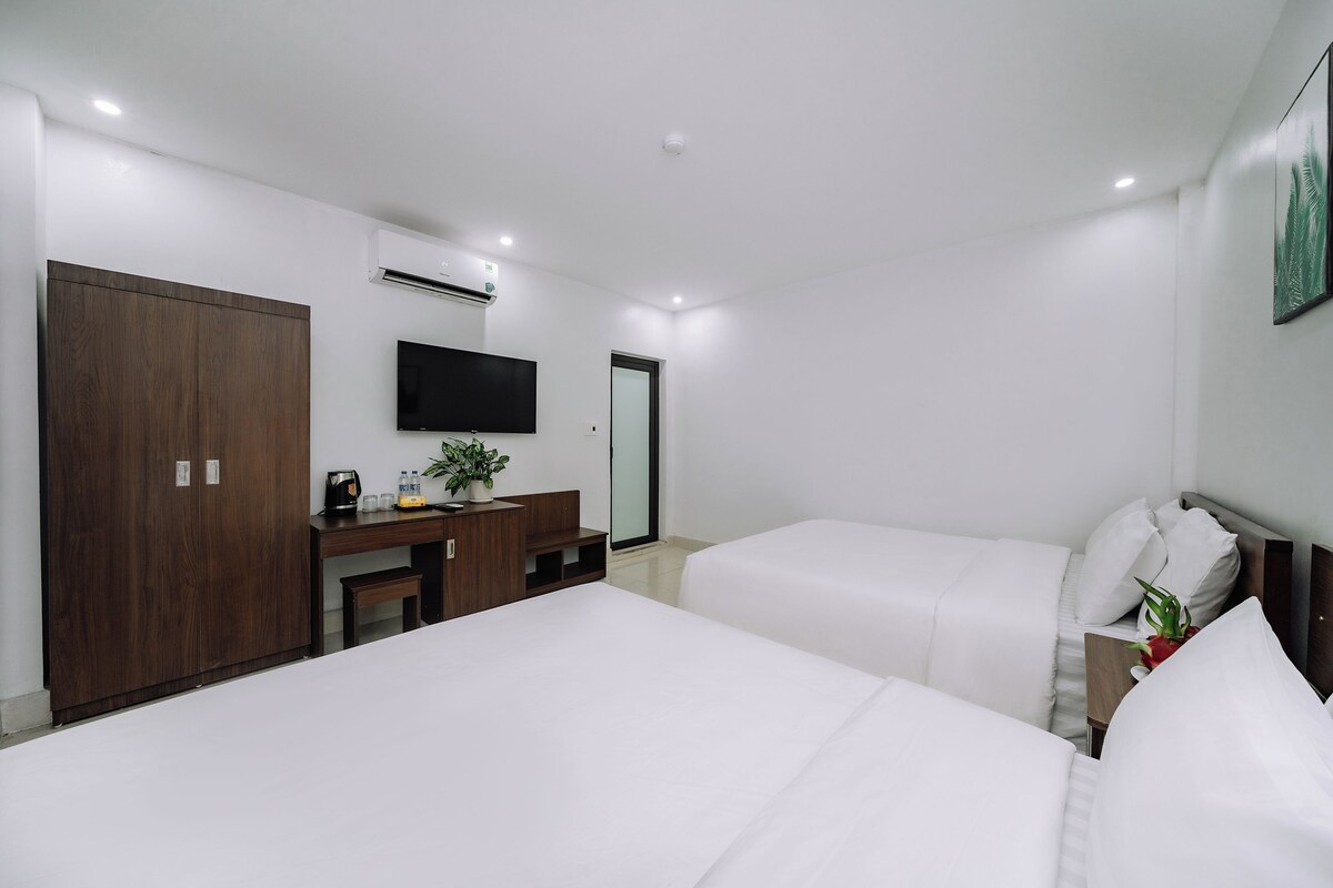 A hotel room with two beds, a desk with a flat-screen TV, a wardrobe, and an air conditioning unit.
