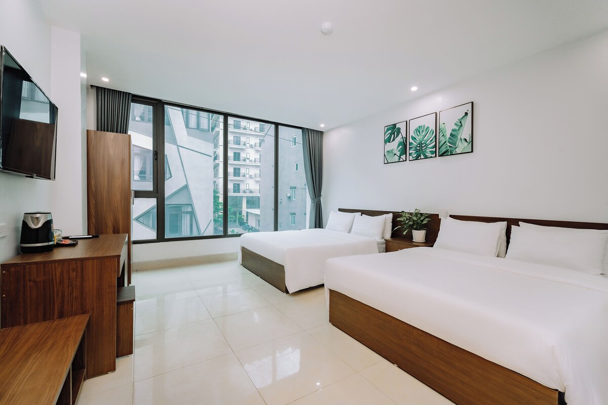 A hotel room with two beds, a desk, a TV, and large windows with a city view.