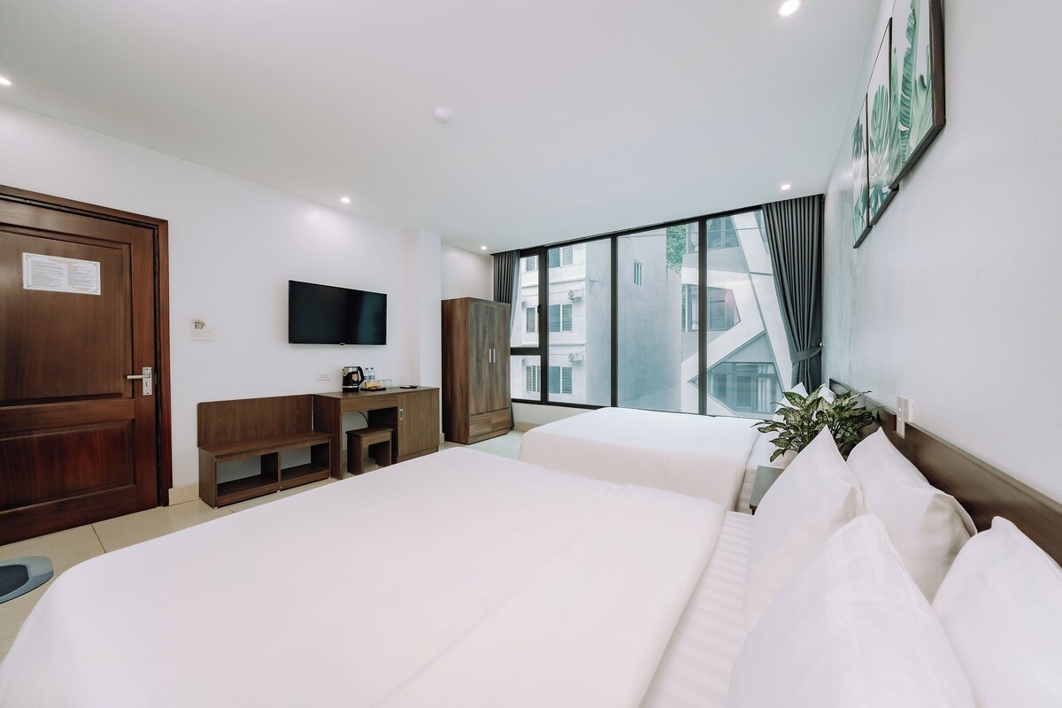 A hotel room with two beds, a desk, a TV, and a large window with a view of buildings.