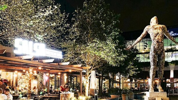 An outdoor dining area with a statue, tables, and chairs, surrounded by greenery and a building with illuminated signage.