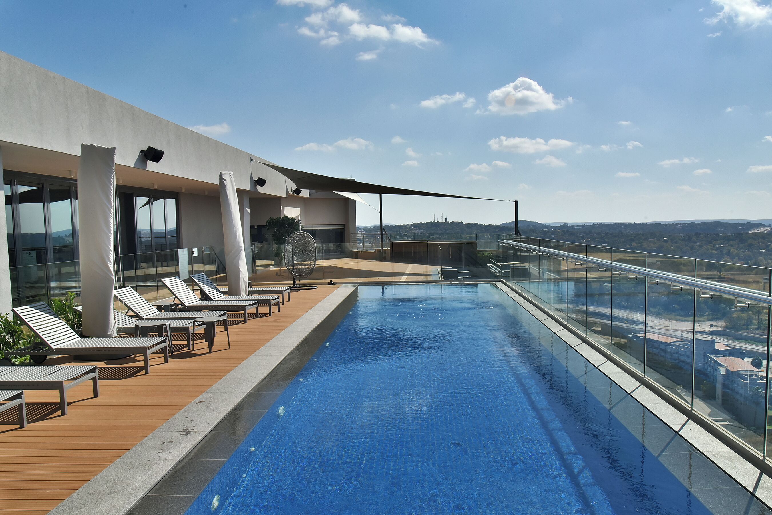 Une piscine à débordement située sur le toit, dotée de chaises longues et d’une balustrade en verre offrant une vue panoramique sur la ville.