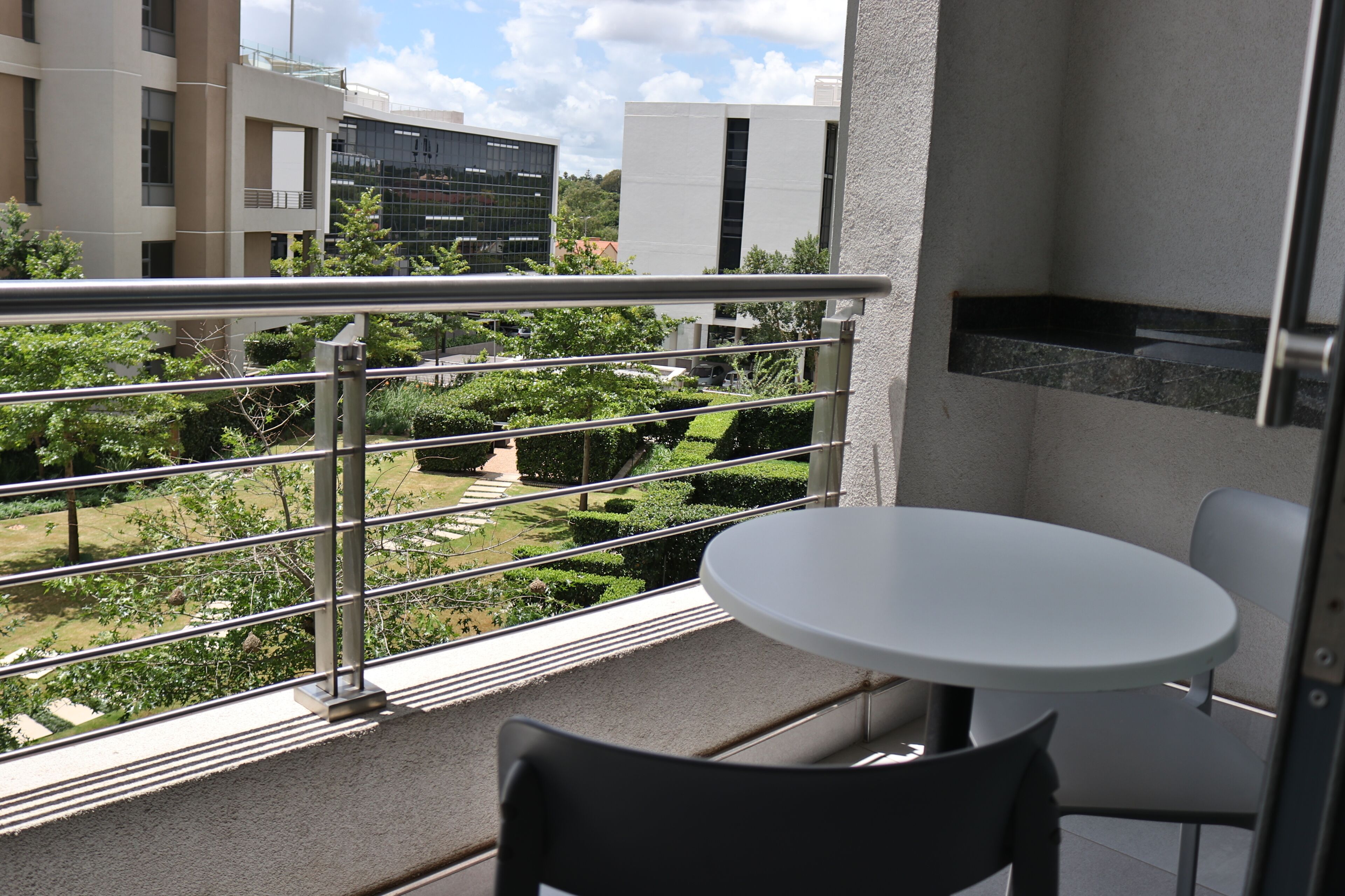 Un balcon avec une table et des chaises blanches, une rambarde en métal et une vue sur un jardin aménagé et des bâtiments en arrière-plan.