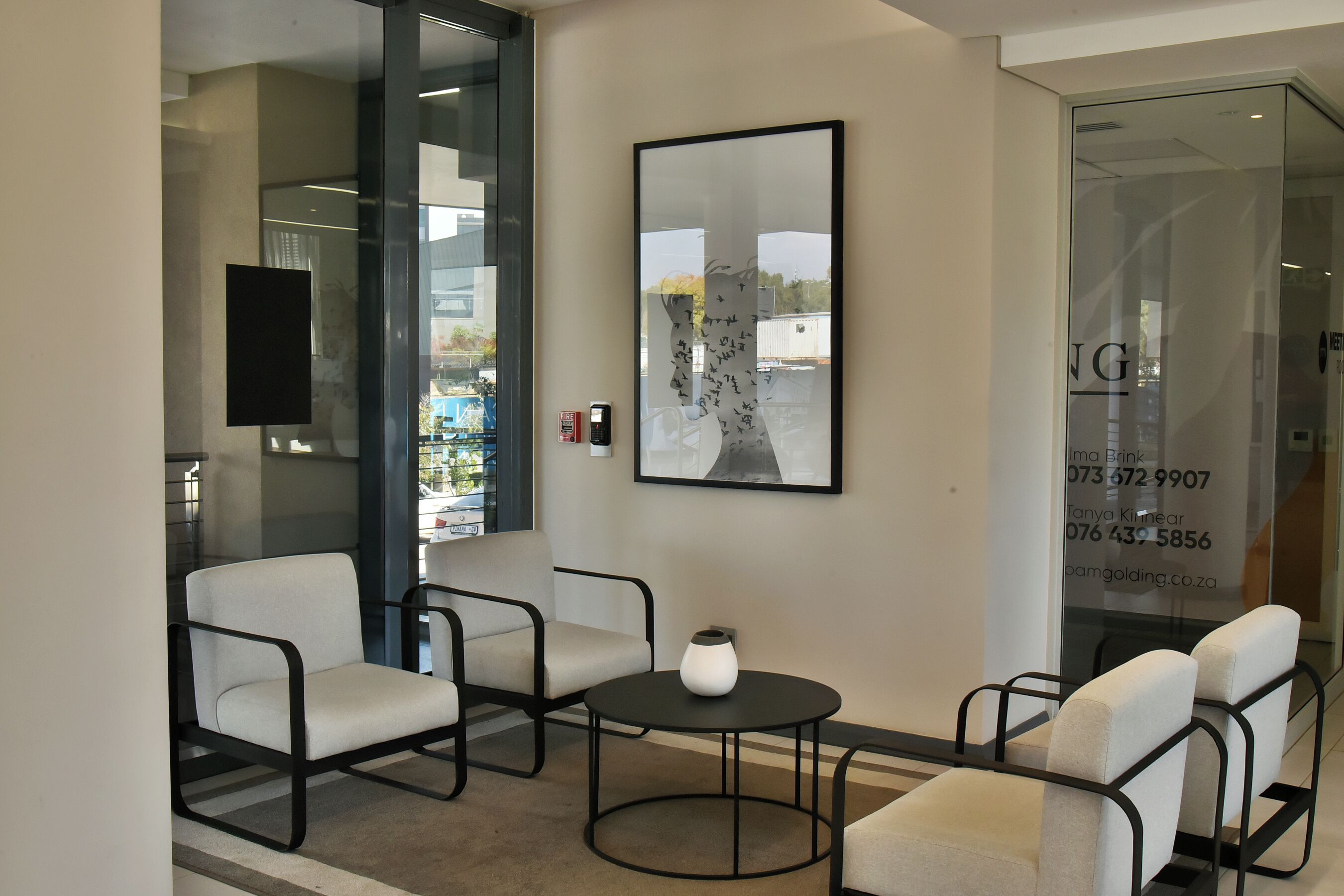 A modern waiting area with a glass wall, a framed artwork, and a contact number on the wall.