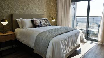 A modern hotel room with a large bed, two bedside lamps, a geometric patterned wall, and a balcony with a city view.