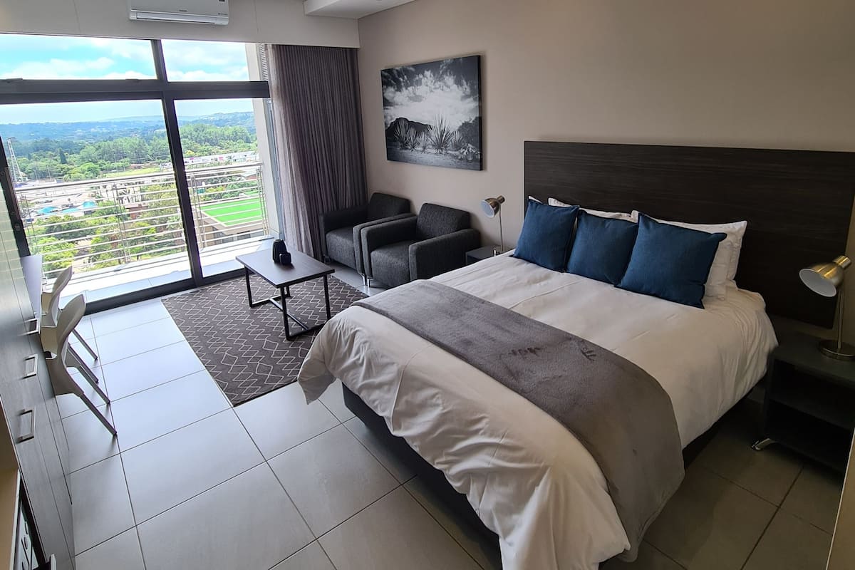 A modern hotel room with a large bed, a seating area with a table, and a view of the landscape through large windows.