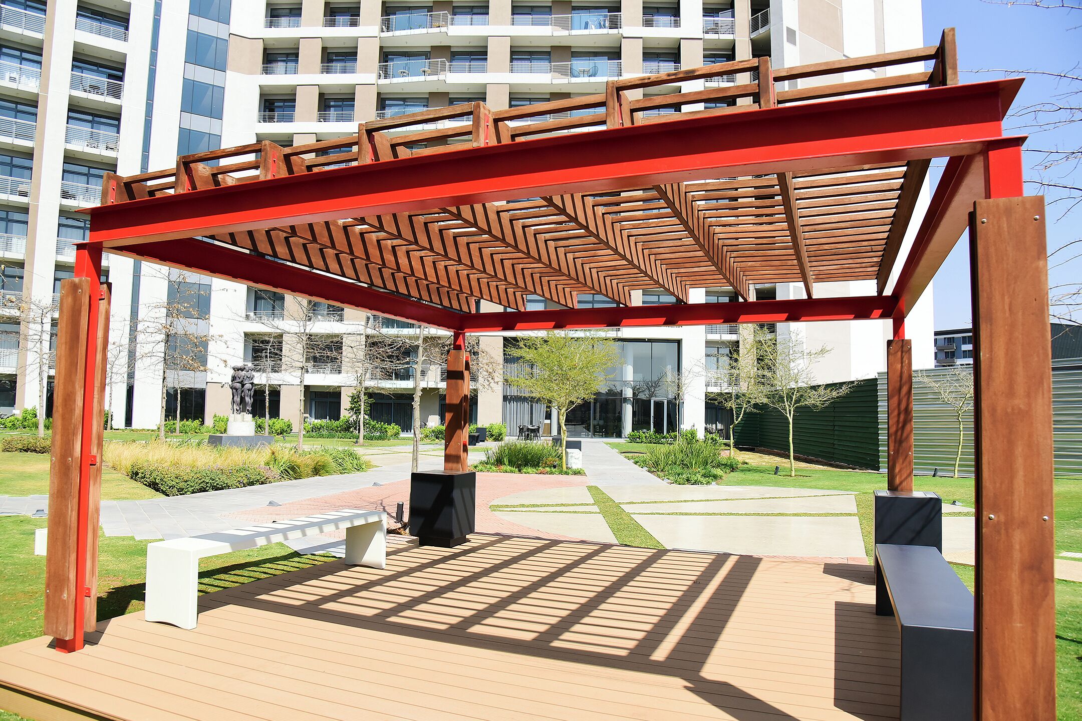 A wooden pergola structure with benches in an outdoor area with a modern building in the background.