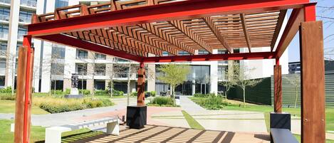 A wooden pergola structure with benches in an outdoor area with a modern building in the background.